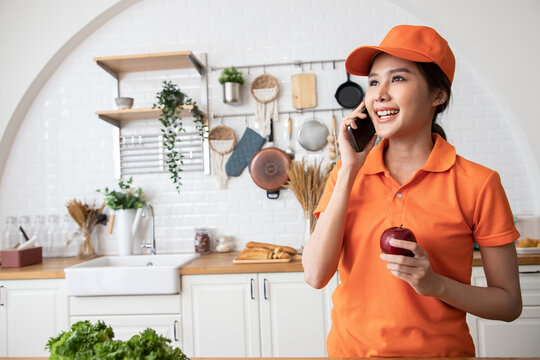 Young Asian caucasian delivery woman in orange uniform talking on the smartphone and holding apple in the kitchen. Checking ecommerce shipping online retail to sell at home