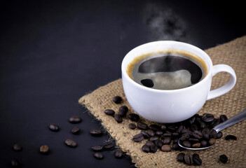 High angle view of a white ceramic cup of black hot Americano coffee with steam on a sackcloth with a pile of roasted coffee beans on black background.