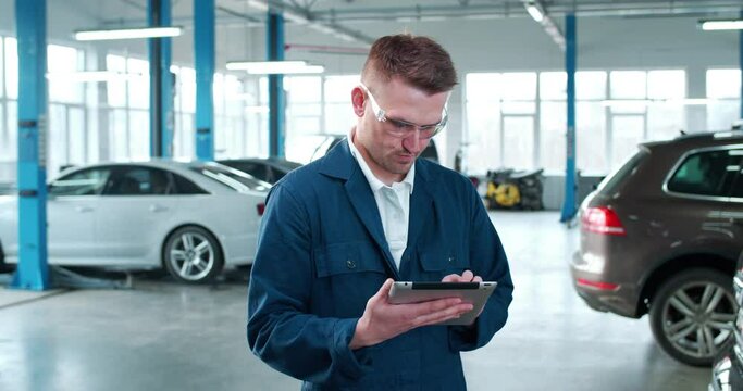 Handsome Caucasian young man in uniform and goggles tapping, texting and scrolling on computer in auto repair service. Male mechanic using tablet device. Workday. Work concept.
