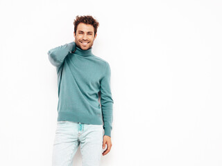 Portrait of handsome smiling model. Sexy stylish man dressed in blue sweater and jeans. Fashion hipster male with curly hairstyle posing near white wall in studio. Isolated