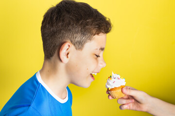 Close-up of a boy in blue, eyeing a cupcake in someone's hand, with a plain yellow backdrop.