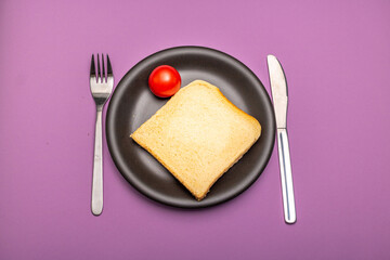 A single tomato and a slice of toast on a plain black plate and cutlery with signs of use against a purple background
