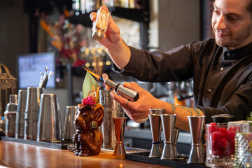 Happy barman sprinkling cinnamon powder and burning tiki cocktail in bar