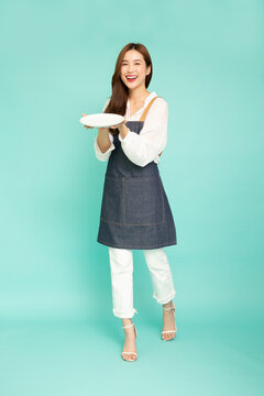 Young Asian Woman Housewife Wearing Kitchen Apron Cooking And Holding Empty White Plate Or Dish Isolated On Green Background, Full Length Composition