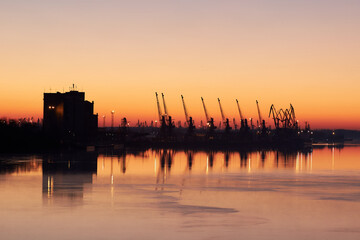 Fototapeta premium River landscape with silhouette of cranes in cargo port at sunrise on quiet surface of autumn Danube river