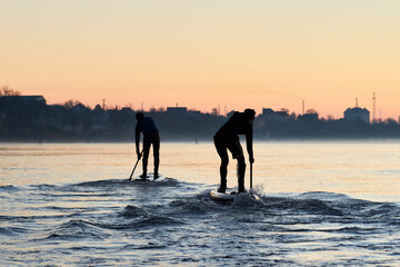 Silhouettes of a two men rowing on stand up paddle boarding (SUP) at sunrise on quiet surface of autumn Danube river. Morning training on SUP