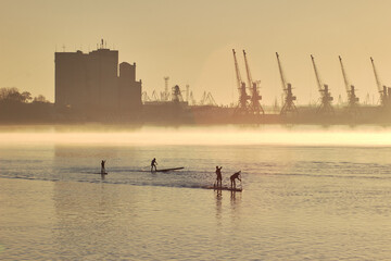 River landscape with silhouettes of people paddle on stand up paddle board (SUP) at sunrise on calm...