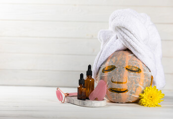 Beauty concept. Cute pumpkin with sheet mask and face care products. Composition on white texture wood. Side view. Copy space