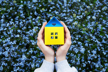 Blue, yellow paper house model in hands. Ukraine flag colors. Refugee, social housing, help support build Ukraine, desire to return home concept. Selective focus. Copy space