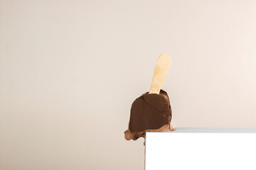 Melting and broken chocolate ice cream bar with stick on white podium. Geometric podium with chocolate popsicle.  Summer creative food concept with copy space.