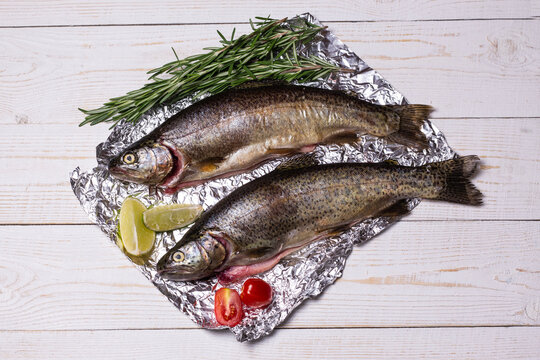 Raw fish, trout with on aluminum foil with ingredients for cooking. Fish, pepper, garlics, tomatoes, lemon and rosemary on white wooden table. Healthy food and dieting concept flat lay, 