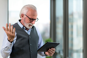 Senior businessman wearing eyeglasses with tablet computer standing close to window in office. Concept of executive using his tablet in his office