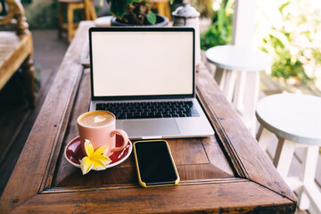 Selective focus on cup of aroma cappuccino with flower standing at cafe table near blank smartphone and blurred laptop with copy space area for your coffee advertising, technology with mockup screens