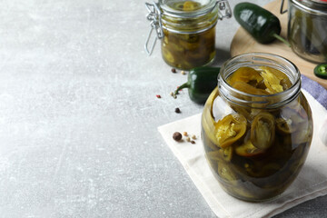 Glass jar with slices of pickled green jalapeno peppers on light grey table, space for text