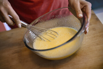 Woman whisking eggs for crepe batter in glass bowl on wooden table, closeup