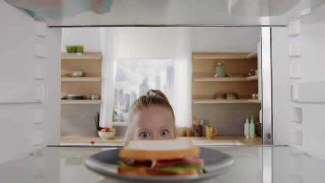 Pretty caucasian girl opens fridge door and taking out tasty sandwich. Little hungry girl looking for food in an empty fridge. POV from inside the refrigerator.