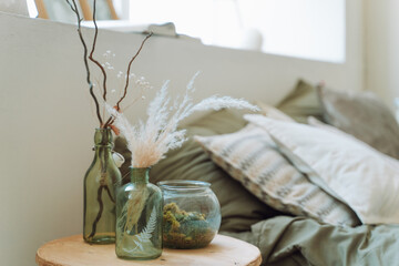 Interior of bright modern bedroom with bed covered with grey bed linen. Different green glass bottles with dry pampas, twigs, decorative plant, round glass vase with moss on beige bedside table. 