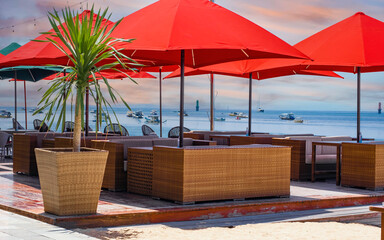 Sea side cafe with wicker furniture and bright red canvas umbrellas and a potted palm tree. Beautiful view of the ocean with boats scattered in distance. Early morning pink and blue sky.  No people