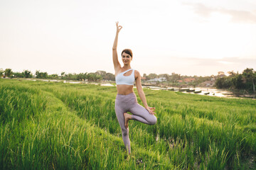 Full length portrait of Caucasian yogi getting nature energy during morning meditation at rice fields in Thailand, sportive female standing in tree pose and practicing hatha yoga during retreat