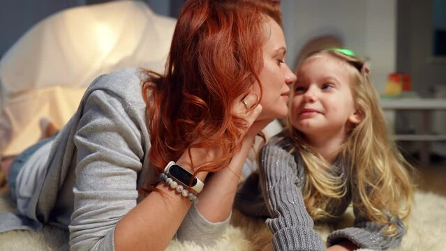 Happy woman kissing nose of cute little girl talking with child indoors. Portrait of positive Caucasian mother and daughter having fun laughing enjoying weekend at tent in living room