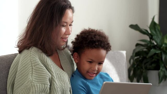 Mixed races family concept. Pretty asian mom and her cute african son sitting on comfort couch indoors. Looking at tablet screen, watching cartoons together , making wide beaming smile. 4k footage