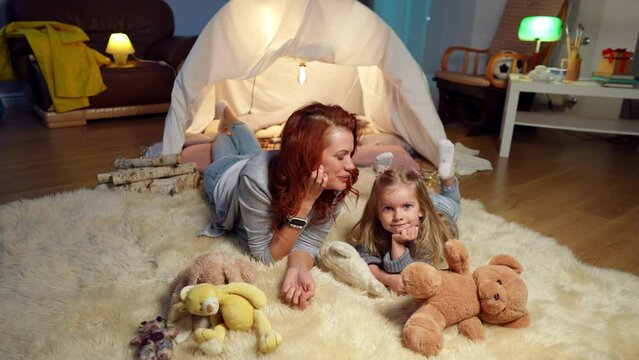 Wide Shot Joyful Mother And Daughter Lying On Carpet At Tent In Living Room Laughing Kissing Each Other. High Angle View Portrait Of Relaxed Cheerful Caucasian Woman And Girl Enjoying Leisure At Home