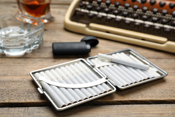 Open case with tobacco filter cigarettes and lighter near vintage typewriter on wooden table, closeup