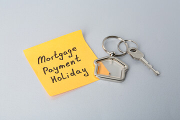 Key with trinket in shape of house and phrase Mortgage payment holiday written on sticky note against light grey background