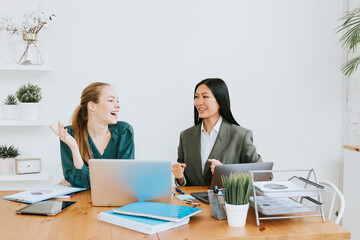 Obraz premium two young business women professionals in formal wear clothes work in modern office using laptop, tablet, brainstorm and search for solutions together, confident independent Asian girl solves problems