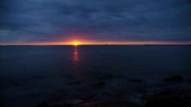 Time lapse of dramatic orange sunset over seascape ocean with clouds moving fast in dark blue sky and stones in water. Composition with long exposures