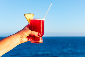 Hand mit Cocktail an der Reling eines Schiffes, blauer Ozean und Himmel im Hintergrund.