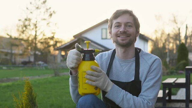 Portrait of smiling man gardener in apron holding insecticide sprayer. Seasonal work in garden.