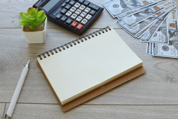 Top view of an open notebook with blank pages, banknotes money calculator and a plant in a white pot on a wooden table background. Flat lay, creative workspace. Business education