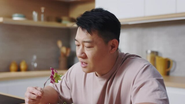 Portrait of sad Asian man sitting at table, holding fork in hand, trying to eat salad in cozy kitchen. Unhappy vegetarian guy. Facial expressions, healthy lifestyle. Indoors. Cooking. Diet concept.