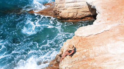 Beautiful girl sitting on a high rock and looking out to sea. top view. Girl in Hat on the edge of cliff. blue sea and high cliffs. Seascape of Cyprus. landscape. beach background. Blue sea, rocks