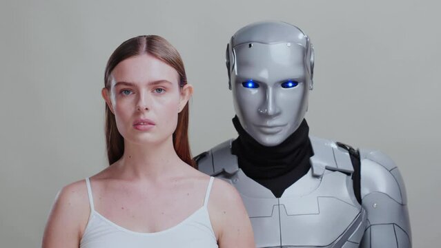 Portrait couple of young caucasian woman and robotic cyborg with blue eyes posing for grey background. Artificial intelligence. Human interaction with robots.