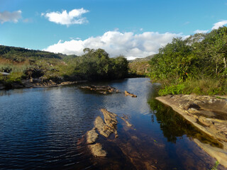 Fototapeta premium Pequeno curso d'água entre lindas pedras e águas escuras, com muita vegetação em volta e céu azul e nuvens, localizada na região rural de Três Barras, município do Serro, Minas Gerais, Brasil.