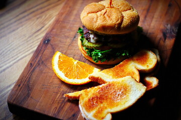 Deluxe Burgers, Hamburgers are being decorated before serving on a wooden cutting board consisting of toasted bread and orange slices.