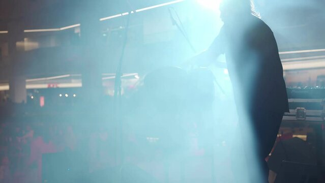 Man with ponytail dreadlocks standing back to camera playing ethnic drums on stage in smoke and spotlights. performance takes place in the lobby of the shopping mall. Rear view medium shoot 