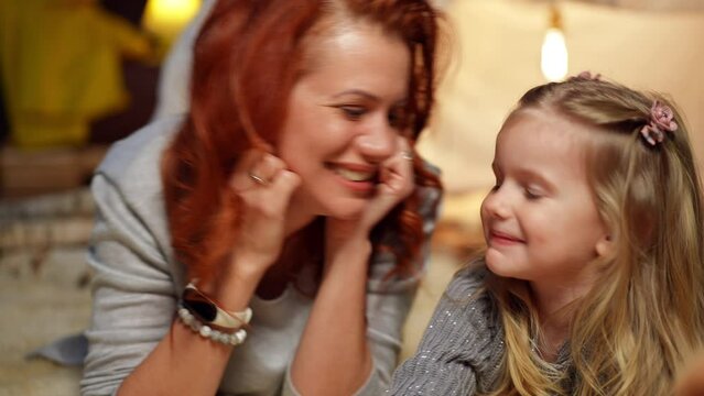 Front view portrait of beautiful mother kissing nose of cute daughter laughing. Happy Caucasian woman enjoying leisure with pretty carefree girl at tent in living room at home indoors
