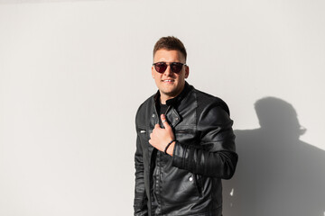 Smiling brutal fashion man with fancy black sunglasses and a trendy black leather jacket outside near a gray wall on a sunny day