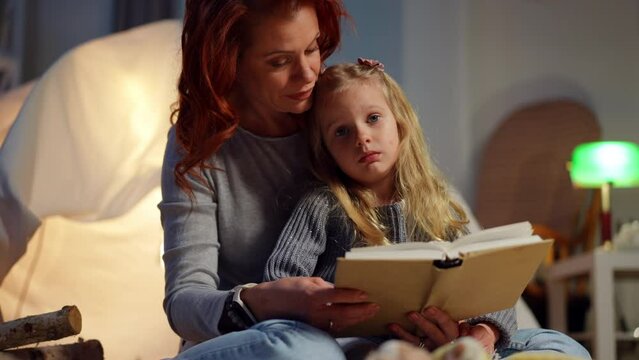 Portrait of cute girl listening to woman reading fairytale at bedtime at tent in living room. Calm Caucasian daughter enjoying leisure with mother at home at night. Family and childhood concept