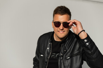 Happy handsome young man with hair in a fancy leather black jacket looks through fashion sunglasses at the camera and smiles against the gray concrete background