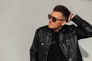 Cool fashion handsome man with hairstyle in black leather jacket and black t-shirt with trendy sunglasses stands and poses near a gray wall on the street
