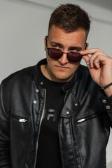 Fototapeta premium Handsome fashionable man in fashion black clothes with a leather jacket looks through sunglasses in the city
