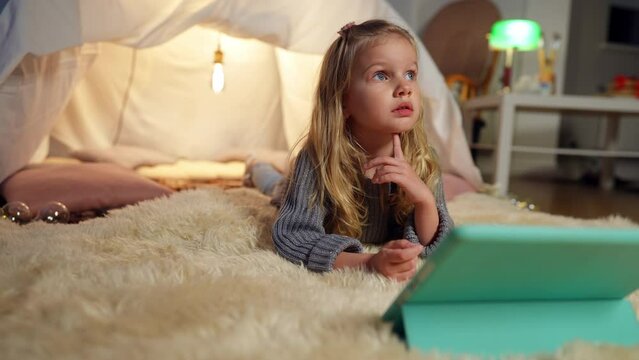 Charming beautiful little girl talking looking up lying with tablet at tent in living room. Wide shot portrait of curios carefree happy Caucasian child resting at home indoors watching cartoons