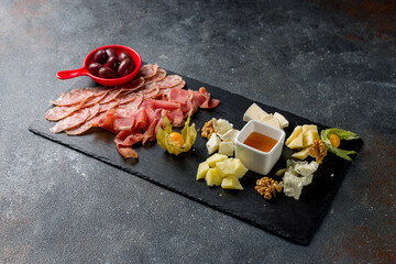 slicing of different meat and different types of european cheese on dark stone plate with olives and honey