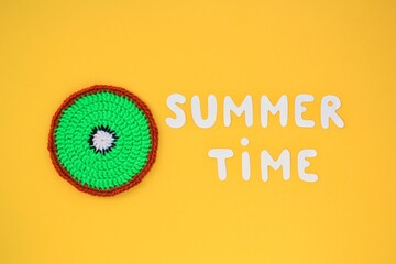 Colourful crochet handmade slice of kiwi and lettering Summertime on yellow background. Hello Summer vacation creative tropical bright minimal food concept. Flatlay, top view holiday composition