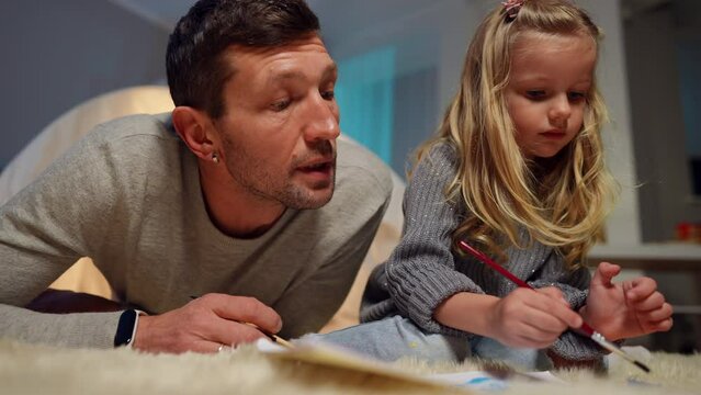 Cute little daughter painting in living room as handsome father talking endorsing child. Portrait of confident Caucasian girl enjoying hobby resting with curios man indoors