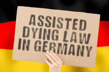 The phrase " Assisted dying law in Germany " on a banner in men's hands with a blurred Deutsch flag in the background. Pain. Care. Clinic. Sickness. Ethical. Ethic. Depression. Drug. End. Judge